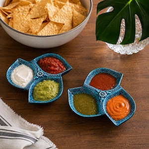 Puede incluir: Dos cuencos de servicio de cerámica azul cerceta con tres secciones, patrones en relieve y un motivo floral central. Cada cuenco contiene tres salsas distintas, incluyendo salsas blancas, rojas, verdes y naranjas en sus compartimentos.
