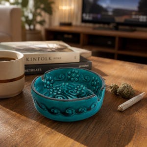 May include: A round, teal ceramic ashtray featuring a raised, embossed pattern of swirling vines and small circular clusters. The ashtray has two notches on the rim for resting items and a smooth, glossy finish with dark accents in the crevices.