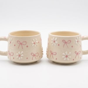 May include: Two cream-colored ceramic mugs featuring a raised, embossed pattern of white daisies, pink bows, and small pink dots. Each mug has a rounded handle and a slightly tapered base, showcasing a consistent repeating design across the exterior.