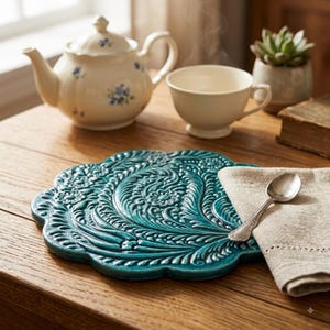 Puede incluir: Un salvamanteles redondo de cerámica color verde azulado con un intrincado patrón floral y de hojas en relieve y bordes festoneados. Una cuchara de metal plateado descansa sobre una servilleta de lino beige doblada sobre el salvamanteles.