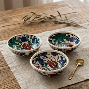 May include: Three small ceramic bowls with a cream-colored base and a hand-painted floral pattern in shades of deep red, blue, and green. Each bowl features a rounded shape and a smooth rim. A small, tarnished brass-colored metal spoon sits nearby.