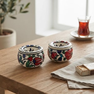Pode incluir: Dois cinzeiros redondos de cerâmica com um padrão floral pintado à mão em vermelho, azul e verde sobre um fundo branco. Cada cinzeiro tem uma borda espessa e curva e uma bacia central profunda, exibindo motivos botânicos detalhados.