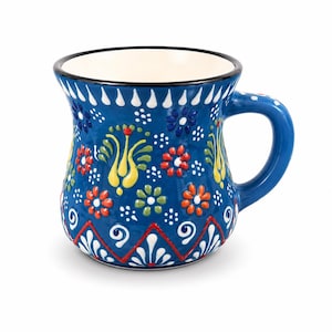 May include: A ceramic mug with a deep blue base color, featuring a hand-painted floral pattern in red, yellow, and green. The design includes white dotted accents and a geometric zigzag border near the base. The interior of the mug is cream-colored.