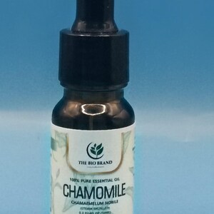Puede incluir: Un pequeño frasco cuentagotas de vidrio oscuro que contiene 10 ml de aceite esencial de manzanilla 100% puro. La etiqueta blanca presenta el texto THE BIO BRAND, CHAMOMILE, CHAMAEMELUM NOBILE, STEAM DISTILLED y 0.3 FLUID OZ (10ML) con un logo de hoja verde.