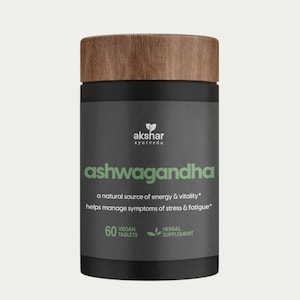 May include: A cylindrical supplement bottle with a dark gray label and a wood-grain textured lid. The label features the text "akshar ayurveda", "ashwagandha", "a natural source of energy & vitality", "60 vegan tablets", and "herbal supplement".