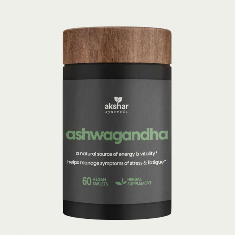 May include: A cylindrical supplement bottle with a dark gray label and a wood-grain textured lid. The label features the text "akshar ayurveda", "ashwagandha", "a natural source of energy & vitality", "60 vegan tablets", and "herbal supplement".