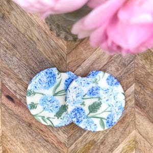 May include: Two circular car cup holder coasters featuring a white background with a repeating pattern of blue hydrangea flower clusters and green leaves. Each coaster has a small semi-circular notch along the top edge for easy removal.