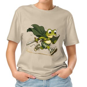 Puede incluir: Una camiseta beige de manga corta con una ilustración gráfica de una rana verde con capa y pantalones cortos de color verde oscuro, corriendo mientras sostiene una espada y un libro abierto. La rana tiene una expresión decidida y está en movimiento.