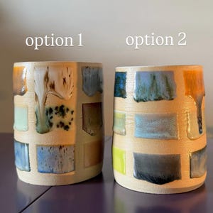 May include: Two ceramic mugs with a beige base and colourful, rectangular glaze patches. "Option 1" features earth tones, while "Option 2" has blues, greens, and yellows. The glaze drips down the sides, adding texture. The mugs are cylindrical.