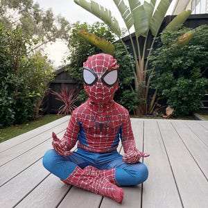 May include: A child wearing a Spider-Man costume, featuring a red and blue design with web patterns. The costume includes a mask with white eye coverings. The child is sitting cross-legged on a wooden deck, with hands in a meditative pose.