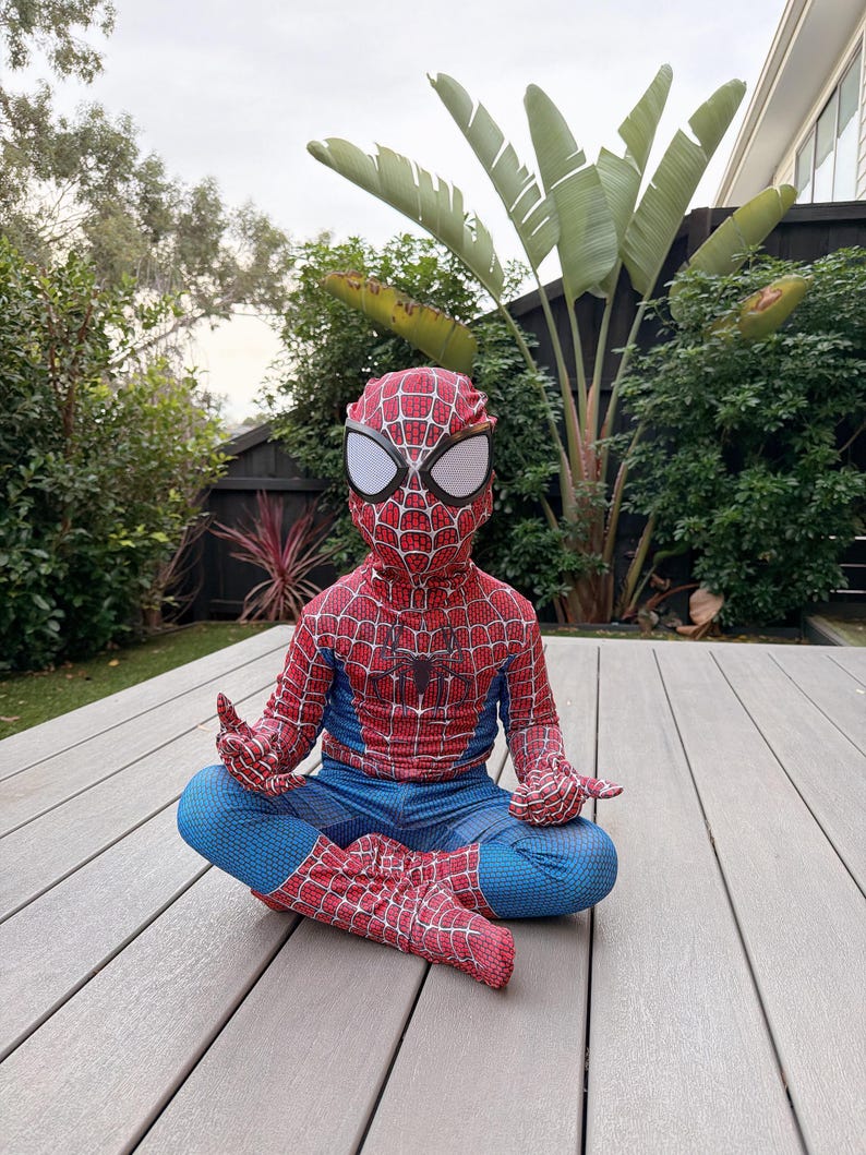 May include: A child wearing a Spider-Man costume, featuring a red and blue design with web patterns. The costume includes a mask with white eye coverings. The child is sitting cross-legged on a wooden deck, with hands in a meditative pose.