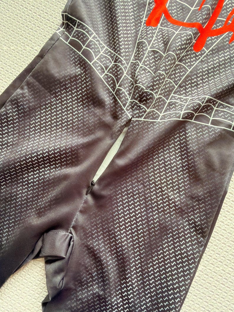 May include: Black jumpsuit with a white web design and red accents. The fabric has a subtle chevron pattern. The jumpsuit appears to be a costume or cosplay outfit.