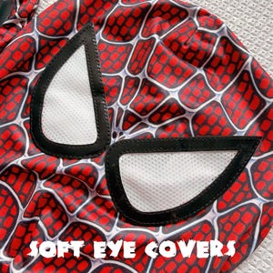 May include: A close-up of a red and black Spider-Man mask with white eye covers. The mask features a web pattern design. The text "SOFT EYE COVERS" is printed in white across the bottom of the image.