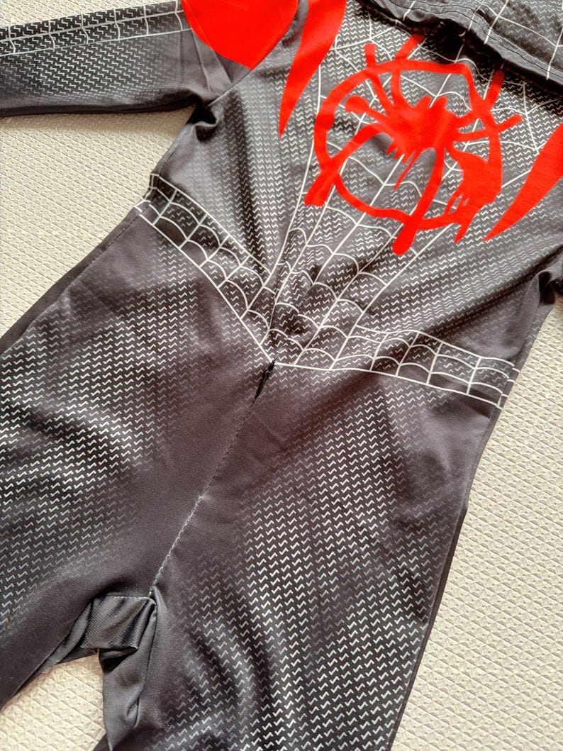 May include: Black and gray jumpsuit with a spider web design and a red spider emblem on the back. The jumpsuit has a textured pattern and a hood. The design is reminiscent of a superhero costume.