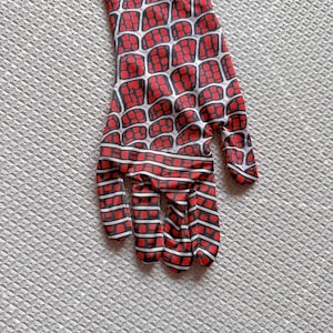 May include: A red and black patterned glove, featuring a web-like design. The glove has a white grid pattern over a red background, with black outlines. The fingers are also red and black striped. The glove is likely part of a costume.