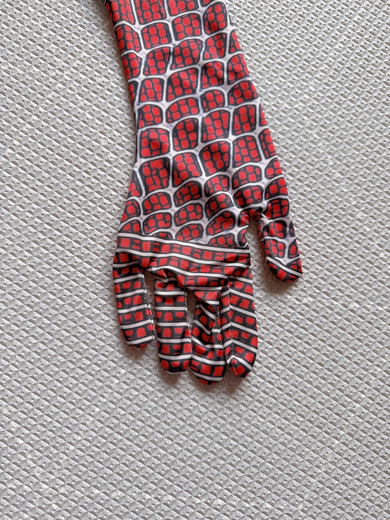 May include: A red and black patterned glove, featuring a web-like design. The glove has a white grid pattern over a red background, with black outlines. The fingers are also red and black striped. The glove is likely part of a costume.