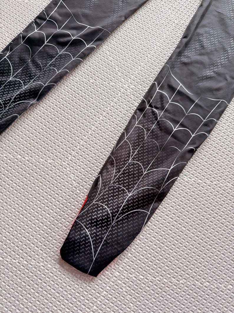 May include: Black leggings with a white spiderweb design. The leggings have a red accent at the bottom and a textured pattern throughout. The spiderweb design is on the front of the leggings.