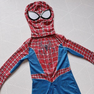 May include: A red, blue, and white Spider-Man costume. The hooded jumpsuit features a web pattern, a spider emblem on the chest, and large white eye shapes on the mask. The costume is designed for children.