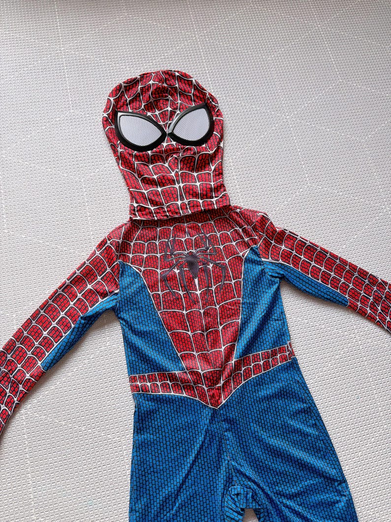 May include: A red, blue, and white Spider-Man costume. The hooded jumpsuit features a web pattern, a spider emblem on the chest, and large white eye shapes on the mask. The costume is designed for children.