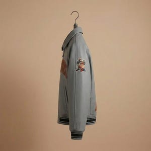 May include: A light gray varsity jacket featuring a fold-down collar, long sleeves with ribbed cuffs, and a front zipper closure. The jacket displays a colorful embroidered patch on the upper sleeve and large orange lettering on the chest area.