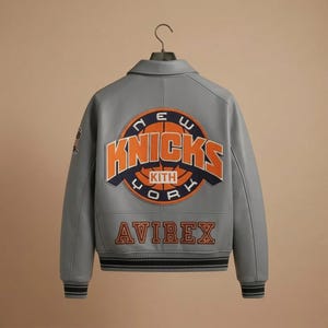 May include: A grey leather varsity jacket featuring a large circular New York Knicks logo on the back with orange and navy blue embroidery. The word AVIREX is embroidered in orange below the logo. The jacket has a collar and striped ribbed cuffs.
