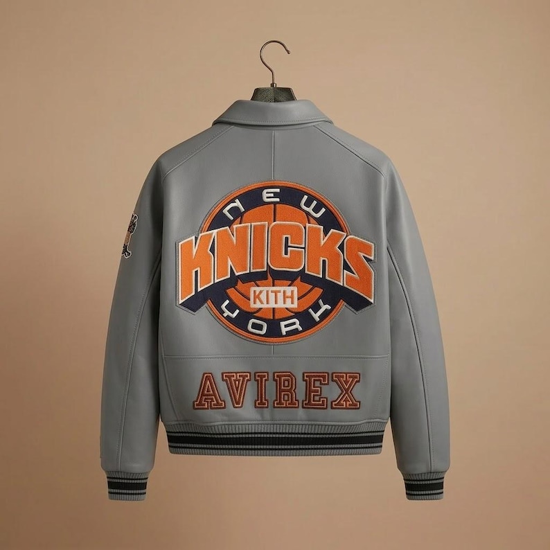 May include: A grey leather varsity jacket featuring a large circular New York Knicks logo on the back with orange and navy blue embroidery. The word AVIREX is embroidered in orange below the logo. The jacket has a collar and striped ribbed cuffs.