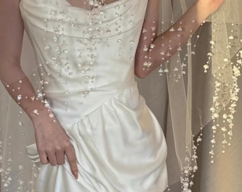 Puede incluir: Un vestido palabra de honor en color marfil con cuerpo drapeado y falda fruncida, acompañado de un velo transparente adornado con aplicaciones florales blancas en cascada y pequeñas perlas que caen sobre los hombros y el frente.