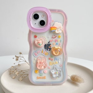 May include: A clear phone case with a wavy pink and purple border. The case is decorated with various 3D charms, including a heart, a bear, a bunny, and the words "Smile", "Happy Day", "Hi Rich!", and "nice".