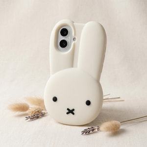 May include: A cream-colored silicone phone case shaped like a rabbit head with two long ears extending from the top. The face features two small black circular eyes and a black cross-shaped mouth. A cutout at the top accommodates a dual-lens camera.