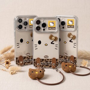 Op de afbeelding: Drie transparante telefoonhoesjes met een Hello Kitty-ontwerp. Elke hoes heeft een bruine strik, zwarte ogen en snorharen. De hoesjes hebben een bruin bandje en een bijpassende Hello Kitty pluche bedel. De tekst "hello kitty" en "free" zijn zichtbaar.