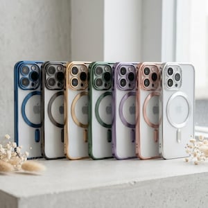 Op de afbeelding: Een rij van zeven doorschijnende smartphonehoesjes met metallic gekleurde randen in blauw, zwart, goud, groen, paars, roségoud en zilver. Elk hoesje is voorzien van een cirkelvormige magnetische oplaadring aan de achterzijde.