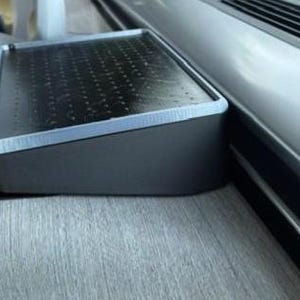 Puede incluir: Un objeto rectangular gris oscuro con una parte superior perforada y un borde plateado. El objeto está en ángulo y parece ser un componente de una estructura más grande, posiblemente un interior de automóvil o un mueble.