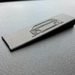 Könnte beinhalten: Ein dunkelgraues, rechteckiges, auto-förmiges Emblem mit abgeschrägter Kante. Das Emblem zeigt eine detaillierte Umrisszeichnung eines Autos, die seine Räder und Karosserie zeigt. Das Objekt befindet sich auf einer strukturierten, dunkelgrauen Oberfläche.