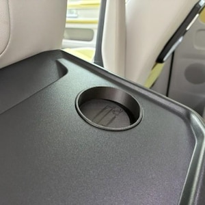 Op de afbeelding: Een donkergrijze autotray met een ingebouwde bekerhouder. De tray is rechthoekig met afgeronde randen en is gepositioneerd tussen twee autostoelen. Het interieur van de auto is zichtbaar op de achtergrond.