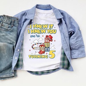 Op de afbeelding: Een wit t-shirt met de tekst I KNEW IT I KNEW YOU we're TURNING 5, voorzien van afbeeldingen van een rode cowboyhoed, alfabetblokken, een kralenarmband en rode cowboylaarzen. Het t-shirt wordt gedragen onder een blauw overhemd en een blauwe spijkerbroek.