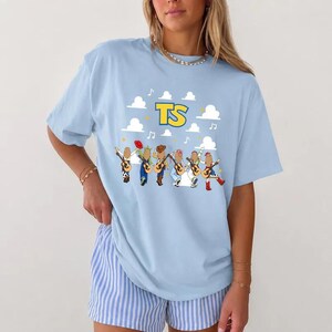 May include: A light blue short-sleeve t-shirt featuring a graphic print of six cartoon figures playing acoustic guitars. Above the figures, the yellow text TS is displayed against a background of white clouds and small musical notes.