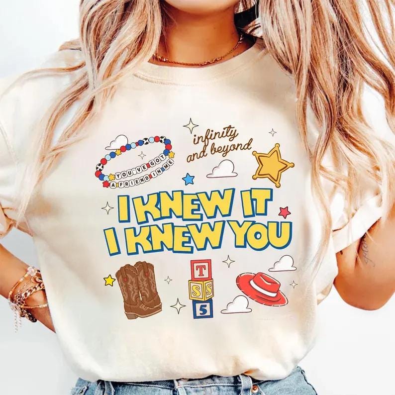 May include: A cream-colored t-shirt featuring a graphic print with the text "I KNEW IT I KNEW YOU" in blue and yellow, "infinity and beyond" in script, a beaded bracelet, a yellow star, brown cowboy boots, alphabet blocks, and a red hat.