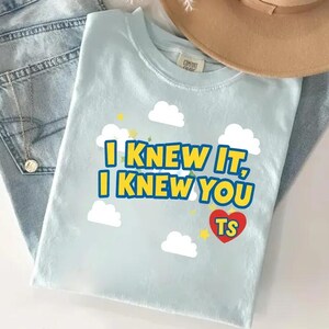 K&ouml;nnte beinhalten: Ein hellblaues T-Shirt mit einem Grafikdruck aus wei&szlig;en Wolken, gelben Sternen und dem Text "I KNEW IT, I KNEW YOU" in fetten blauen Buchstaben. Unter dem Text befindet sich ein rotes Herz mit den gelben Buchstaben "TS" in fetter Schrift.