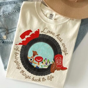 May include: A cream-colored t-shirt featuring a graphic of a black tire framing a scene with cartoon characters. The text reads Love has ways of bringing things back to life. A red cowboy hat and a red cowboy boot are positioned near the tire graphic.