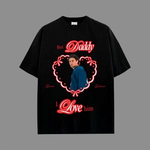 May include: Black t-shirt featuring a heart-shaped design with the words "But Daddy I Love him" in red script. The design includes a photo of a person wearing a blue shirt. The names "Garret" and "Graham" are also included.