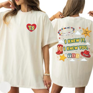May include: An off-white t-shirt featuring a red heart with the letters TS on the front. The back displays a beaded bracelet, a sheriff badge with the text Infinity and Beyond, red cowboy boots, a red hat, and the text I Knew It, I Knew You, TS5.
