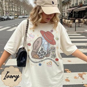 May include: An ivory short-sleeve t-shirt featuring a graphic print of a cow-print cowboy boot, a red hat, and blocks with the letters T, S, and 13. The boot has the name Taylor printed on the sole. A black shoulder bag with a chain strap is worn.