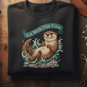 Pode incluir: Camisola preta com um desenho de lontra de desenho animado. A lontra é castanha e bege, relaxando na água com o texto "Go With The Flow" numa faixa. O desenho inclui água, plantas e flores cor-de-rosa.