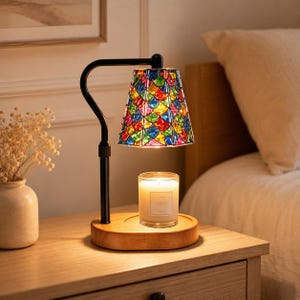 Puede incluir: Una lámpara calentadora de velas con brazo de metal negro y base de madera. La pantalla tiene un diseño colorido estilo vitral con un patrón de escamas repetido en tonos azul, verde, rojo y amarillo. Una vela en vaso transparente está debajo.