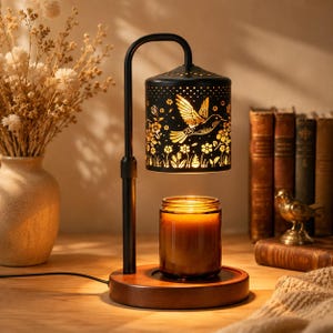 Puede incluir: Una lámpara calentadora de velas de metal negro con una pantalla cilíndrica que presenta diseños recortados de un colibrí y flores. La lámpara tiene una base de madera y se sitúa sobre una vela en frasco de vidrio que emite una luz cálida.