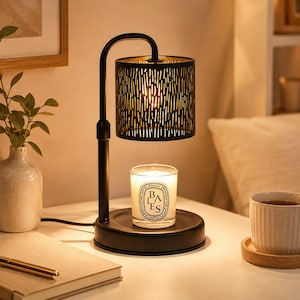 May include: A black metal candle warmer lamp with a cylindrical shade featuring vertical cutouts. The lamp has a curved arm and a circular base holding a glass candle jar with a white label that reads BAIES. The lamp emits a warm glow.
