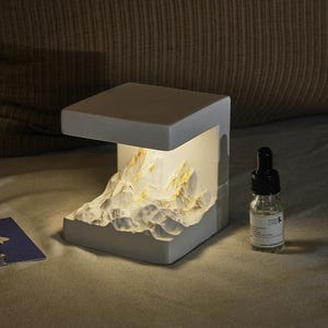 May include: A white cube-shaped lamp featuring an internal sculpted mountain range with gold-coloured accents. The light illuminates the textured peaks from within. Beside the lamp sits a small clear glass bottle with a black dropper cap.