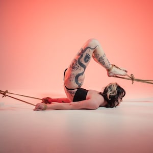 May include: A person with extensive black ink tattoos on their legs, wearing a black top, is positioned on the floor. Their wrists and ankles are bound with thick tan rope, which is connected to two large, dark, jagged rocks on either side.