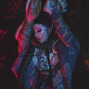 May include: A person with tattoos and makeup is entwined with a snake. The person wears a white top with a corset design and is bound with rope. The background is dark with red and green hues.