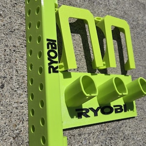 May include: A lime green plastic tool storage mount featuring the black text RYOBI printed on the side and front. The unit includes a vertical pegboard-style side panel with multiple circular holes and a front section with two U-shaped hooks.